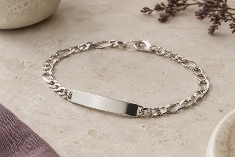 Bracelet argenté à chaîne Figaro et plaque lisse personnalisable posé sur une pierre claire